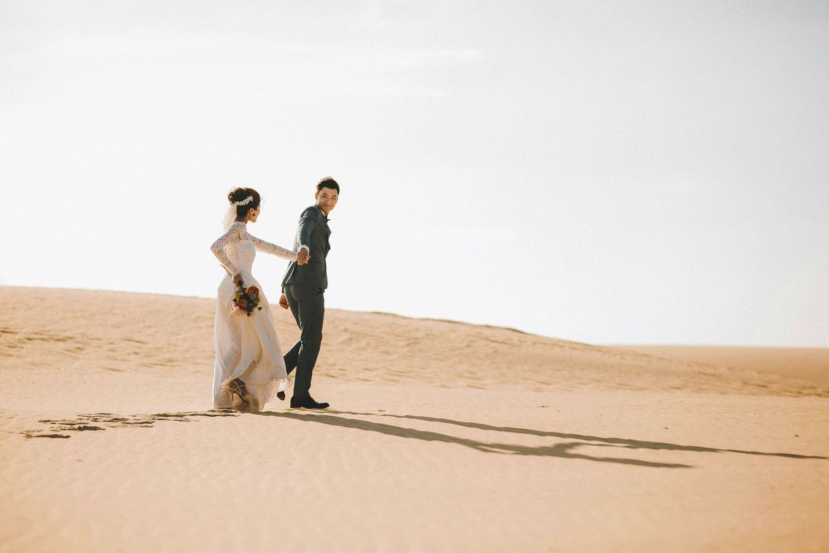 A bride and groom walking hand in hand across sand dunes in warm evening light